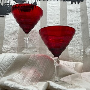 May include: Two red wine glasses with a textured bowl and clear, twisted stem. The glasses are set against a white and gray striped background. The glasses are likely made of glass and are suitable for serving wine.