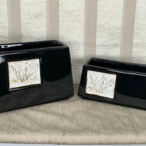 May include: Two black ceramic rectangular planters with white floral designs. The larger planter is taller and wider than the smaller planter.