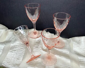 May include: Four pink glass wine glasses with a textured surface. The glasses are on a white fabric background.