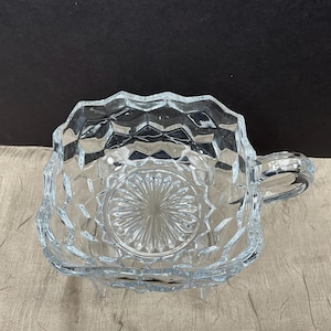 May include: Clear glass serving bowl with a handle. The bowl has a square shape with a scalloped rim and a diamond pattern. The base of the bowl has a circular design. The bowl is likely used for serving sauces or dips.