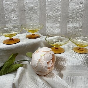 May include: Four vintage dessert glasses with yellow bowls and amber-colored stems and bases. The glasses are arranged on a white, textured surface, with a pink flower and green leaves in the foreground. The glasses have a textured pattern.