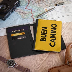 Puede incluir: Un portapasaporte negro con una tarjeta de crédito y un portapasaporte amarillo con el texto "BUEN CAMINO" en negro.