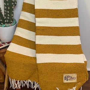 May include: Mustard yellow and white striped blanket with a fringed edge. The blanket is draped over a wooden chair. A tag on the blanket reads "Sedona Blanket". A cactus in a white pot is in the background.
