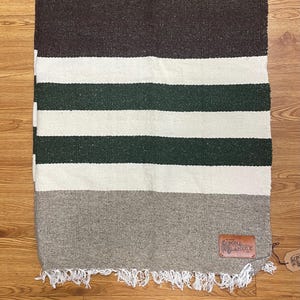 Puede incluir: Una manta rectangular tejida con una sección superior marrón, seguida de rayas blancas y verdes, y una sección inferior gris. La manta tiene un borde con flecos y una etiqueta de cuero que dice "Sedona Blanket".