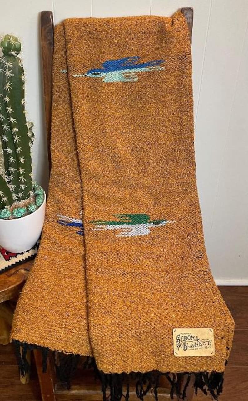 May include: A woven blanket in shades of orange and brown, draped over a wooden chair. The blanket features blue, green, and white embroidered designs. The bottom edge has black fringe, and a label reads "Sedona Blanket."