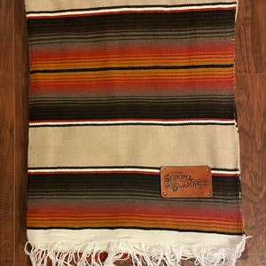 May include: A white and brown striped throw blanket with a fringe border. The blanket has a leather patch with the text "The Original Sedona Blanket Co."