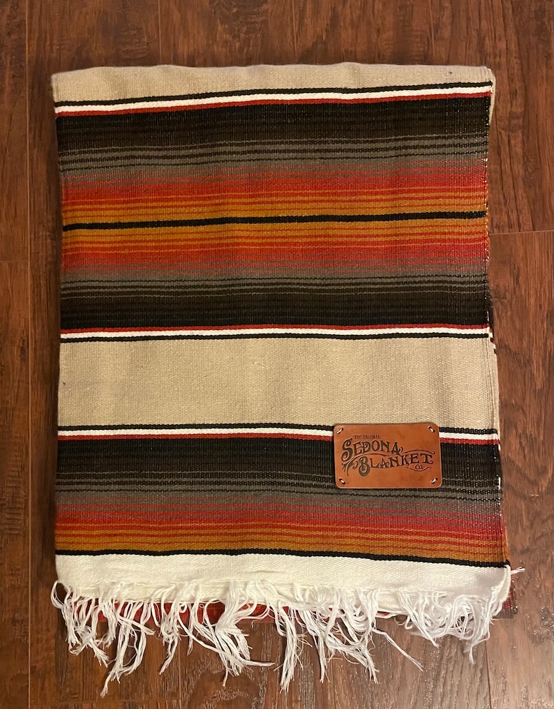 May include: A white and brown striped throw blanket with a fringe border. The blanket has a leather patch with the text "The Original Sedona Blanket Co."