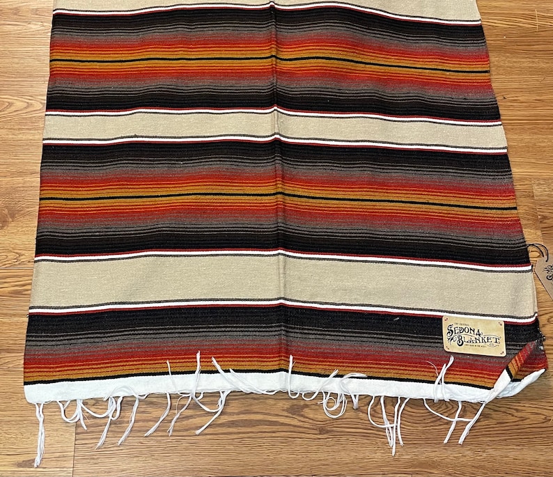May include: A striped woven blanket with a fringe border. The blanket has a brown, red, and white color scheme. A brown leather tag with the text "Sedona Blanket" is attached to the blanket.