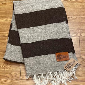 Puede incluir: Una manta doblada con franjas anchas alternas grises y marrón oscuro. La manta tiene un borde con flecos y una etiqueta de cuero que dice "Sedona Blanket". Se adjunta una etiqueta a juego.