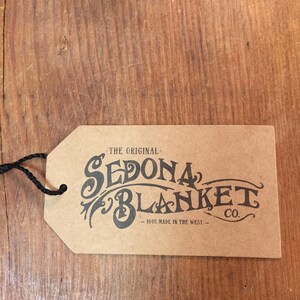 May include: A brown paper tag with the words "SEDONA BLANKET CO." in black script. The tag also reads "THE ORIGINAL" and "100% MADE IN THE WEST." A black string is attached to the tag.