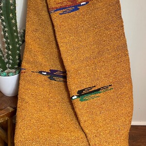 May include: Mustard-colored woven blanket with decorative embroidered accents in blue, red, and green. The blanket has a textured appearance and black fringe along the bottom edge. The blanket is draped over a wooden chair.