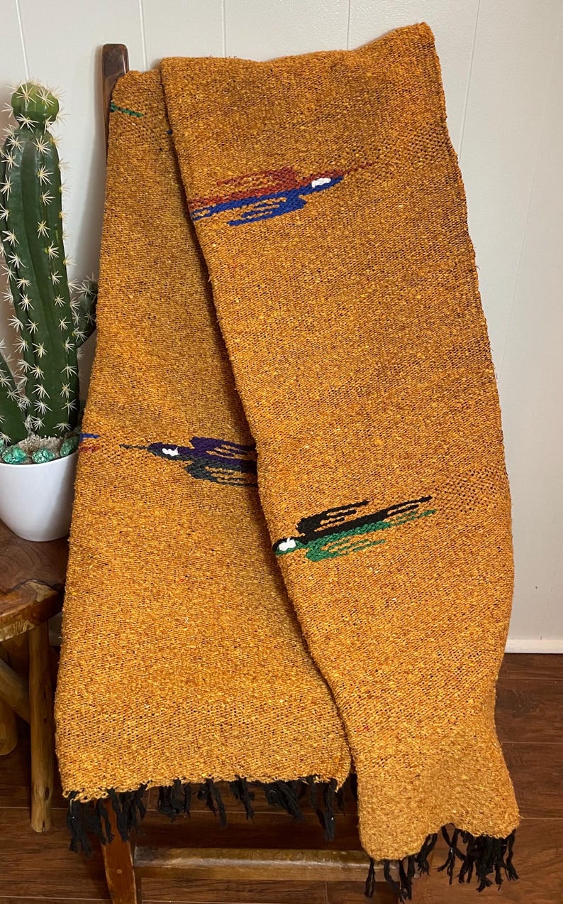 May include: Mustard-colored woven blanket with decorative embroidered accents in blue, red, and green. The blanket has a textured appearance and black fringe along the bottom edge. The blanket is draped over a wooden chair.