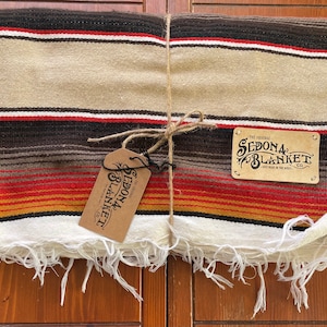 May include: A folded striped blanket with fringe edges, tied with twine. The blanket is beige, brown, red, and yellow. The label reads "The Original Sedona Blanket Co. Hand Made in the West."