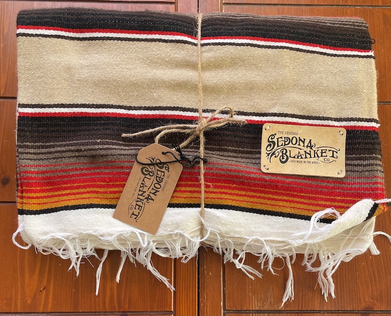 May include: A folded striped blanket with fringe edges, tied with twine. The blanket is beige, brown, red, and yellow. The label reads "The Original Sedona Blanket Co. Hand Made in the West."