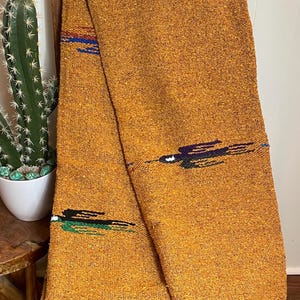 May include: Mustard-colored woven blanket with black fringe. The blanket has a textured appearance and is decorated with horizontal stripes in blue, green, and purple. The blanket is draped over a wooden chair.