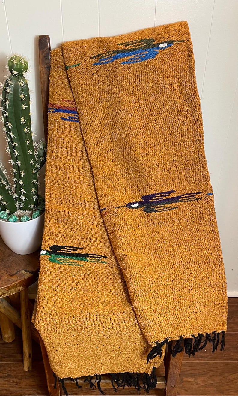 May include: Mustard-colored woven blanket with black fringe. The blanket has a textured appearance and is decorated with horizontal stripes in blue, green, and purple. The blanket is draped over a wooden chair.