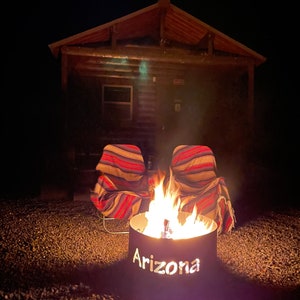 May include: A fire pit with the word "Arizona" cut out of the metal, surrounded by two striped chairs in front of a wooden cabin at night.