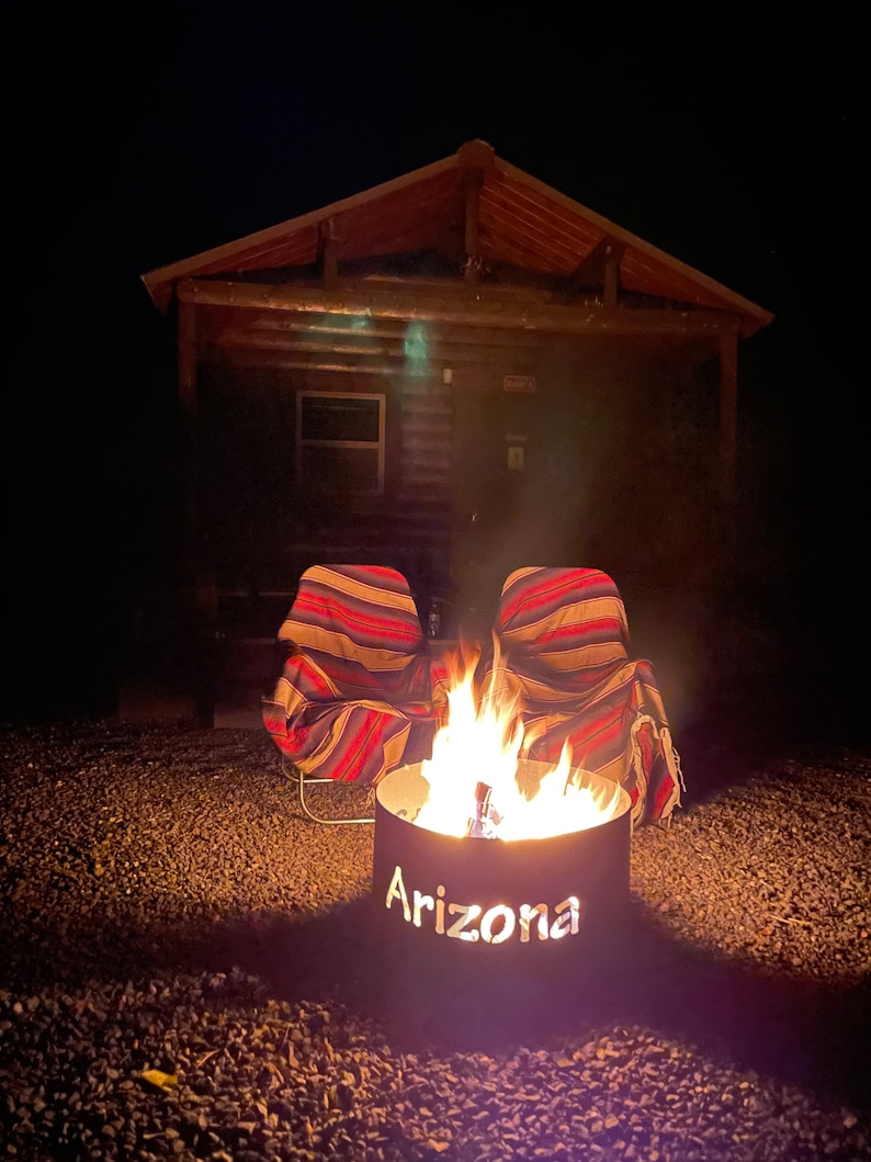 May include: A fire pit with the word "Arizona" cut out of the metal, surrounded by two striped chairs in front of a wooden cabin at night.