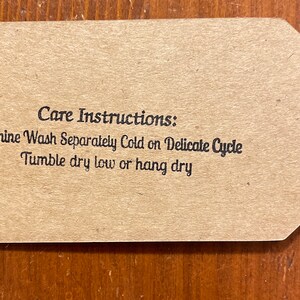 May include: A brown tag with a black string attached. The tag has the words "Care Instructions: Machine Wash Separately Cold on Delicate Cycle, Tumble dry low or hang dry" printed in black.