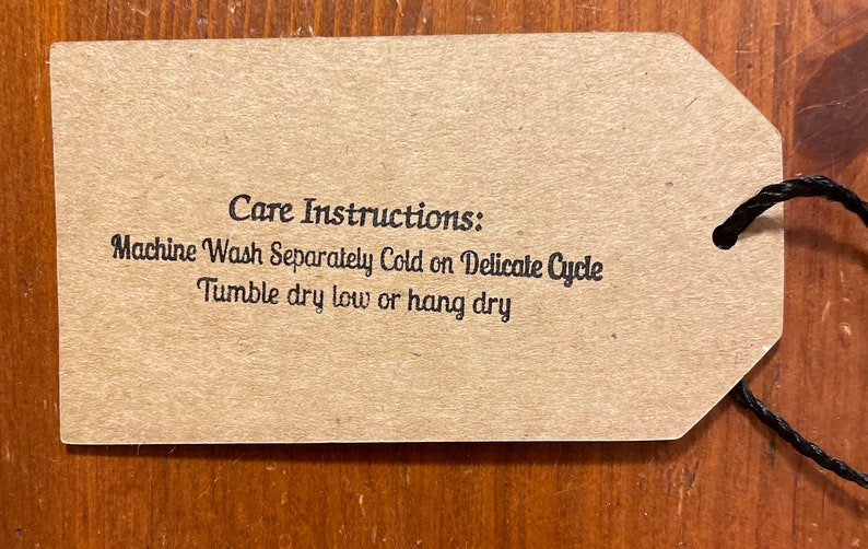 May include: A brown tag with a black string attached. The tag has the words "Care Instructions: Machine Wash Separately Cold on Delicate Cycle, Tumble dry low or hang dry" printed in black.
