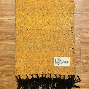 Puede incluir: Una manta tejida de color amarillo mostaza con flecos negros. La manta tiene una etiqueta que dice "The Original Sedona Blanket Co. Hand Made in the USA."