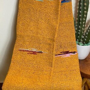May include: Mustard yellow woven blanket with embroidered accents in teal, red, and pink. The blanket is draped over a wooden chair and has a fringed edge. A cactus in a white pot is visible in the background.