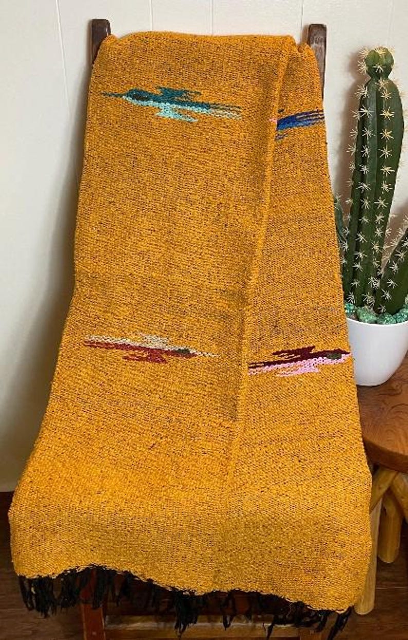 May include: Mustard yellow woven blanket with embroidered accents in teal, red, and pink. The blanket is draped over a wooden chair and has a fringed edge. A cactus in a white pot is visible in the background.
