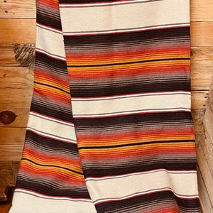 May include: A striped throw blanket with orange, red, brown, and white stripes. The blanket is folded and draped over a wooden chair. The blanket has a label that says "Sedona Blanket Co."
