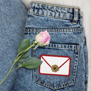 Puede incluir: Primer plano de unos vaqueros azules con un parche decorativo de un sobre blanco con borde rojo y sello de corazón dorado. Una rosa rosa con hojas verdes descansa sobre los vaqueros.
