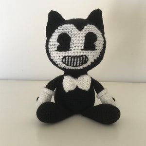 May include: A black and white crocheted plush toy of Bendy, a cartoon character from the video game Bendy and the Ink Machine. The toy is sitting with its legs crossed and has a white bow tie.