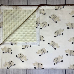 May include: A white baby blanket with a beige and brown rhino pattern. One side of the blanket is a soft, white minky fabric with a subtle dot pattern. The other side is a white cotton fabric with a repeating pattern of beige rhinos wearing brown sweaters.
