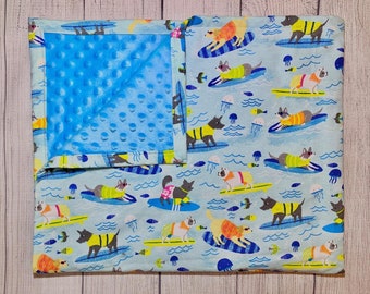 Puede incluir: Una manta para bebé azul y blanca con un lado suave y texturizado y un lado impreso con perros haciendo surf en las olas. El lado impreso presenta un patrón repetitivo de perros con camisas coloridas y gafas de sol, montando tablas de surf sobre un fondo azul con olas blancas y medusas.