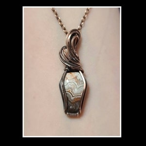 May include: A silver wire wrapped pendant featuring a light brown, white, and gray agate stone. The pendant is attached to a silver chain.