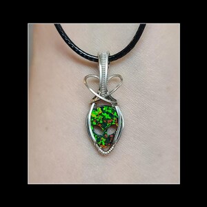 May include: A silver pendant with a black cord necklace. The pendant features a wire-wrapped design around a green and black opal stone carved in the shape of an alien head. The alien head has two eye sockets and a mouth.