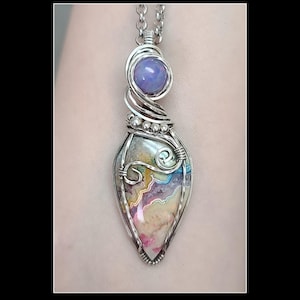 May include: A handcrafted pendant necklace featuring a teardrop-shaped stone with colorful, swirling patterns in pink, yellow, and blue. The stone is encased in a silver wire wrap, with a small, round, purple stone at the top. The pendant hangs from a silver chain.
