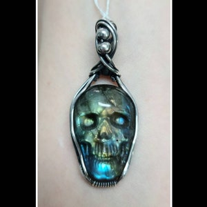 May include: A silver wire wrapped pendant featuring a green, blue, and gold labradorite gemstone carved into a skull shape.