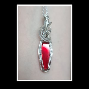 May include: A silver pendant with a rectangular, deep red gemstone. The gemstone is encased in a wire-wrapped design, with intricate swirls at the top. The pendant hangs from a delicate silver chain, creating a striking contrast.