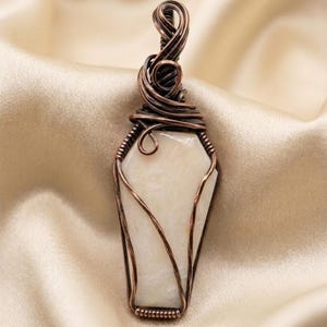 May include: A coffin-shaped pendant featuring a smooth, off-white stone encased in dark, oxidized copper wire wrapping. The wire forms an intricate, swirling bail at the top, with thin strands securing the stone along its tapered edges.