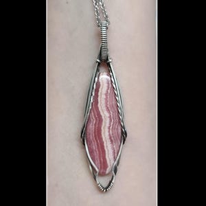 May include: A handcrafted pendant featuring a pink and white striped gemstone, encased in a silver-colored wire frame. The pendant hangs from a delicate silver chain, creating a unique and elegant necklace.