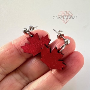 May include: A pair of red maple leaf earrings with silver posts. The earrings are made of wood and have a shiny finish.