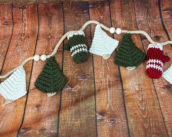 Könnte beinhalten: Eine festliche Häkelgirlande mit abwechselnd weißen, grünen und roten weihnachtlichen Formen. Die Girlande umfasst Weihnachtsbäume, Fäustlinge und Strümpfe, die alle durch ein naturfarbenes Seil mit Holzperlen verbunden sind. Die Girlande wird auf einer Holzoberfläche präsentiert.