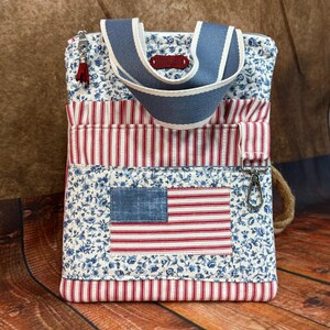 May include: A patriotic tote bag featuring an American flag design. The bag has a blue floral print, red and white stripes, and a blue handle. A small red tassel hangs from the zipper.