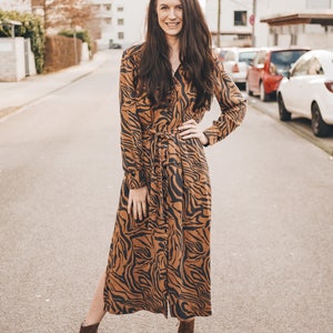 May include: A woman wearing a long brown and black tiger print dress with a long sleeve and a tie belt. She is wearing brown ankle boots and has long brown hair. She is standing on a paved street with cars parked in the background.