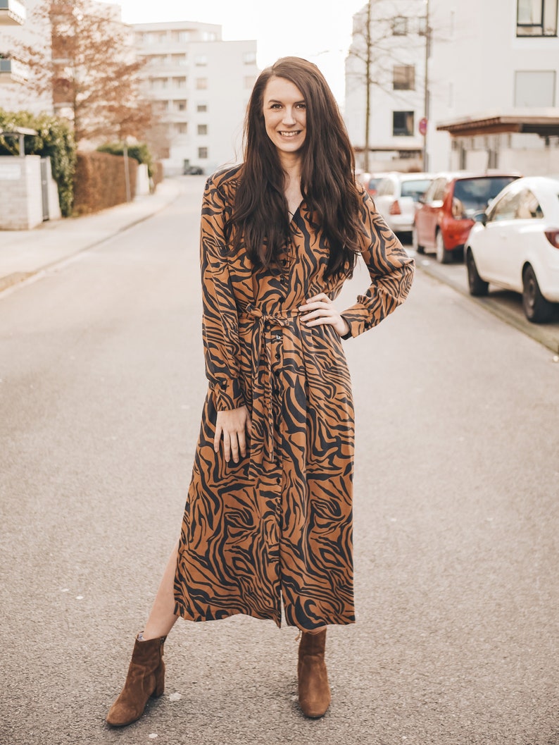 May include: A woman wearing a long brown and black tiger print dress with a long sleeve and a tie belt. She is wearing brown ankle boots and has long brown hair. She is standing on a paved street with cars parked in the background.