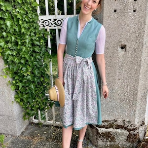 Puede incluir: Una mujer lleva un vestido tradicional b&aacute;varo verde con una blusa blanca y una falda de estampado floral. Lleva un sombrero de paja con una cinta rosa.
