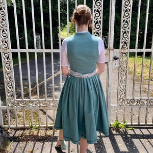Puede incluir: Un vestido de dirndl de color verde azulado con un ribete de encaje blanco y un cintur&oacute;n de cinta con estampado floral. El vestido tiene una falda plisada y lo lleva puesto una persona de pie delante de una puerta de metal blanco.