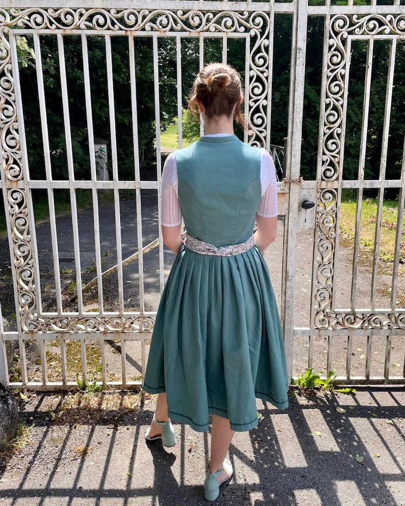 Puede incluir: Un vestido de dirndl de color verde azulado con un ribete de encaje blanco y un cintur&oacute;n de cinta con estampado floral. El vestido tiene una falda plisada y lo lleva puesto una persona de pie delante de una puerta de metal blanco.