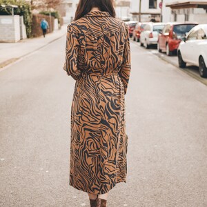 May include: A woman walks down a city street wearing a long brown and black tiger print dress with a belt and brown ankle boots.