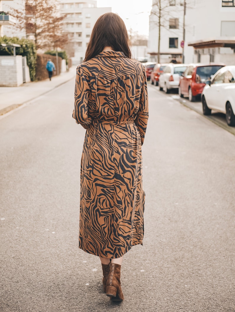 May include: A woman walks down a city street wearing a long brown and black tiger print dress with a belt and brown ankle boots.