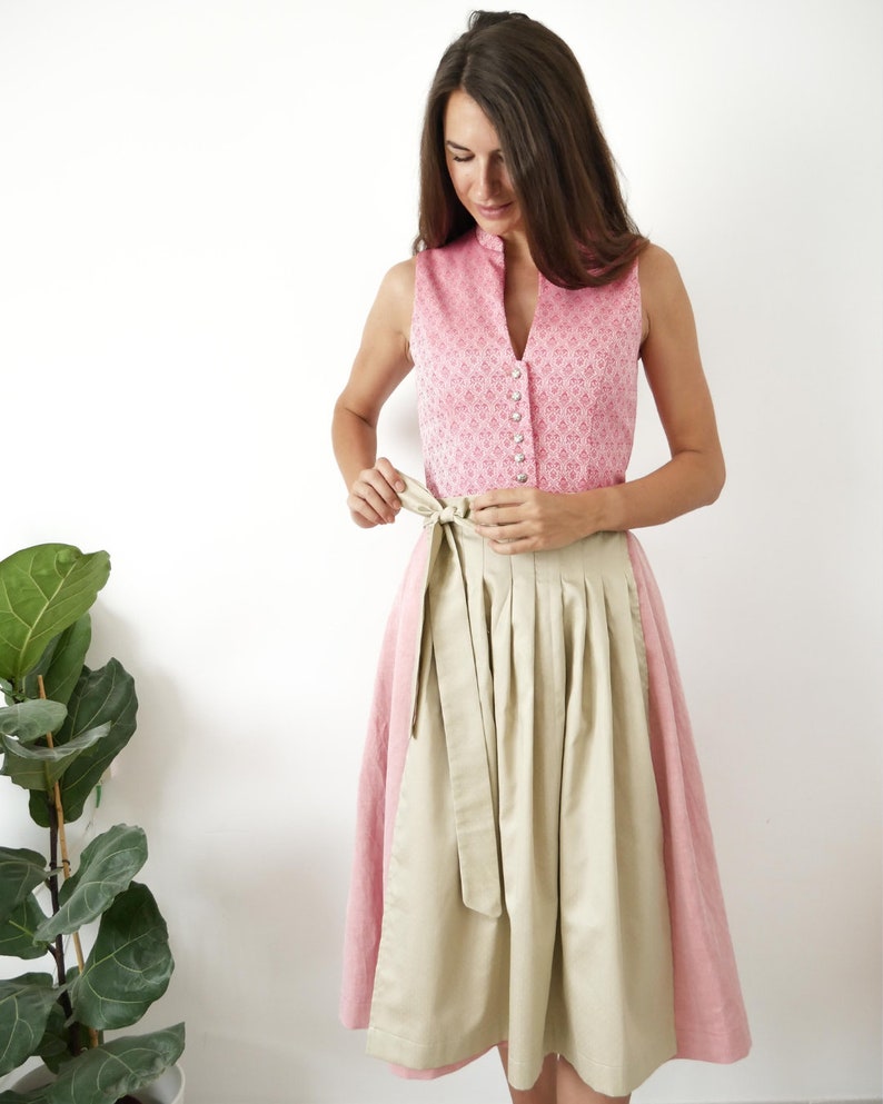 Puede incluir: Un vestido tradicional alem&aacute;n, llamado dirndl, de color rosa y beige con un corpi&ntilde;o blanco con botones y una falda beige. La falda tiene pliegues y un cintur&oacute;n a juego.